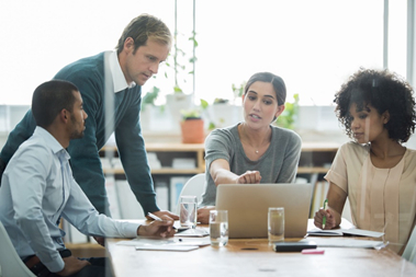 Two men and two women sat and stood over a table in an office with glasses of water on the table looking at a laptop